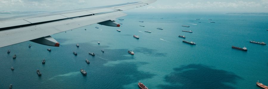 A plane flies over the sea, which is full of transport ships